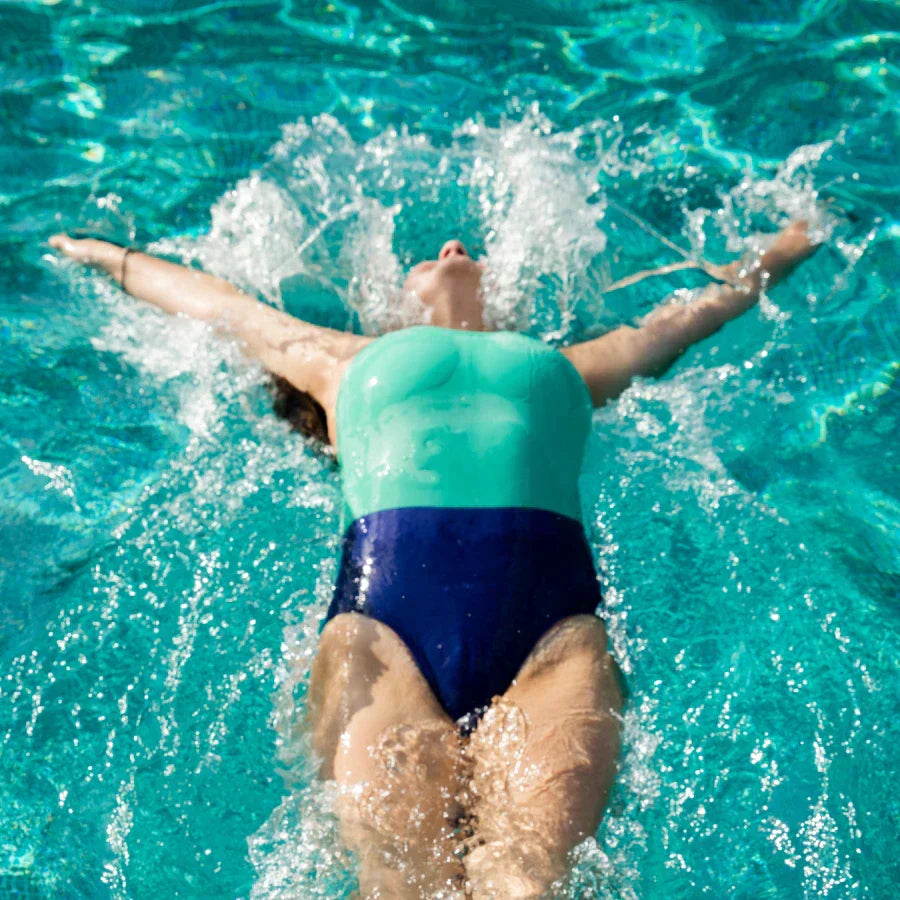High-angle view of a swimmer in a teal and navy one-piece period swimsuit, floating on her back in a bright blue pool. Water splashes around her as she moves.